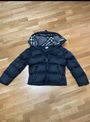 Svart Burberry pufferjacka med huva - Svart pufferjacka från Burberry med avtagbara ärmar, vilket gör den till en väst. Jackan har en huva med klassiskt rutigt Burberry-foder i grått, vitt och rött. Dragkedja framtill och två sidofickor med dragkedja. Märkeslogga på ena ärmen.