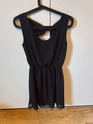 Svart kort klänning med rosett från Love - Supersöt svart kort klänning från Love i storlek XS. Klänningen är ärmlös med markerad midja och har en snygg rosettdetalj och cut-out i ryggen. Tunt, lätt material som ger ett flowy fall. Perfekt för dig som gillar stilrena och feminina detaljer.