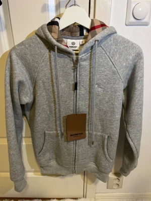 Grå Burberry hoodie  - Grå hoodie från Burberry med klassiskt rutigt foder i huvan och broderad logga på bröstet. Tröjan har dragkedja framtill, snörning i huvan och två fickor. Tillverkad i mjuk bomullsmix, perfekt för chill dagar. Den är helt ny aldrig använt för den är för liten 