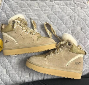 Beige UGGS LOWMEL  - Säljer ett par beige UGG boots med plattformssula och grovt mönstrad yttersula. Skorna har snörning med glittriga guldfärgade skosnören, mjuk päls på insidan och detaljer i mocka och syntet. Perfekta för dig som vill ha både stil och komfort under kalla dagar. Köpta från Australien, har aldrig använts. Men kartongen finns tyvärr inte. Det är storlek 38 på skorna men passar både en storlek uppåt men också neråt. Jag har redan ett par svarta så säljer dessa. Pris kan absolut diskuteras 