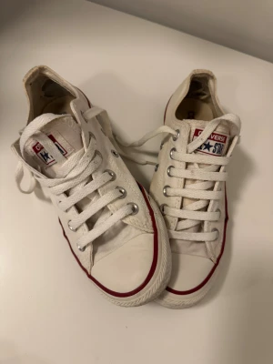 Vita Converse All Star sneakers - Säljer ett par klassiska vita Converse All Star sneakers i storlek 37. Skorna har lågt skaft, rund tå och snörning med metallöglor. Yttermaterialet är i textil och sulan i gummi, med röd rand längs kanten. Perfekt för dig som gillar tidlös streetstil.