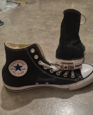 Converse Chuck Taylor All Star High - Svarta Converse Chuck Taylor All Star high-top sneakers med vit gummisula och klassisk rund tå. Skorna har vit snörning, kontrastsydda detaljer och det ikoniska All Star-märket på sidan. Ovandelen är i slitstark canvas och insidan är ljusbeige.