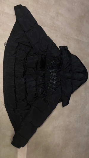 Svart pufferjacka från Trapstar - Svart pufferjacka från Trapstar med huva och snygga detaljer. Jackan har ett stort Trapstar-emblem på insidan och texten 'IT'S A SECRET'. Fylld med syntetmaterial och har dragkedja framtill. Perfekt för kalla dagar och har en clean streetwear-look.