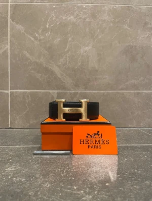 Svart H bälte med guldspänne storlek 115 - Exklusivt svart bälte från H med ikoniskt H-spänne i guldtonad metall. Bältet är tillverkat i äkta skinn och levereras i original orange H box med certifikat. Perfekt accessoar för dig som vill ha en lyxig och stilren look.