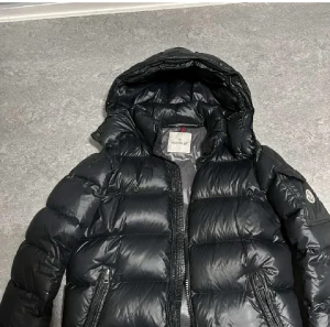 Svart Moncler dunjacka med huva - Säljer en svart dunjacka från Moncler med glansig finish och huva. Jackan har dragkedja framtill, två sidofickor med dragkedja och ett grått foder med serietidningsdetalj. Perfekt för kalla dagar och har en riktigt schysst puffer-look.