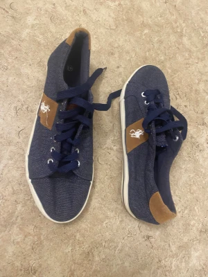 Mörkblå sneakers med mockadetaljer - Snygga mörkblå sneakers med vita sulor och bruna mockadetaljer vid häl och sida. Skorna har snörning och en broderad vit logga på sidan. Ovandelen är i textil och ger en avslappnad men stilren look.