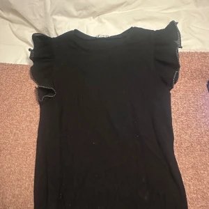 Svart topp med volangärmar - Svart topp med rund hals och snygga volangärmar som har vit kontrastsöm längs kanten. Storlek m men passar s.  Toppen är ribbad och ärmlös, vilket ger en stilren men ändå lekfull look. Perfekt att styla med jeans eller kjol för en trendig outfit.