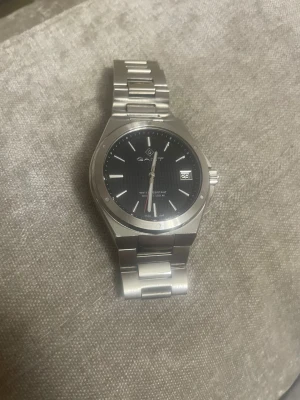 GANT herrklocka med svart urtavla - Snygg herrklocka från GANT med rund svart urtavla och datumvisning. Boetten och länken är i silverfärgat metall, vilket ger en stilren och modern look. Klockan är vattentät upp till 100 meter och har tydliga indexmarkeringar samt en röd sekundvisare.