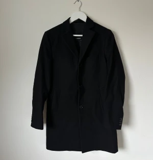 Svart ullrock från J.Lindeberg - Klassisk svart rock från J.Lindeberg i storlek 44. Tillverkad i 80% ull och 20% polyamid med foder i viskos. Rak passform, tre knappar framtill, fickor på sidorna och slits bak. Perfekt för dig som vill ha en stilren och tidlös look. 5O3