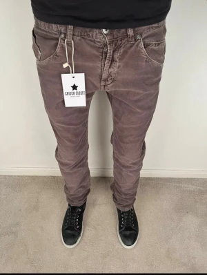 Bruna manchesterbyxor från Dondup - Säljer ett par bruna manchesterbyxor från Dondup med snygga detaljer och röd söm på bakfickan. Byxorna har låg midja, smal passform och klassiska fickor fram och bak. Perfekta för dig som vill ha en chill men ändå stilren look. W27-28, L30. Jag säljer då dom var för små