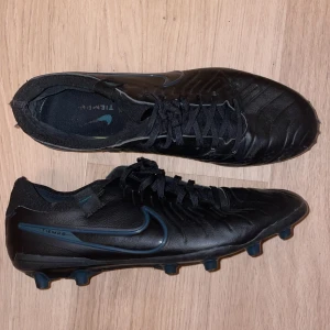 Nike Tiempo svarta fotbollsskor - Svarta Nike Tiempo fotbollsskor med klassisk Swoosh i mörkblått på sidan. Skorna har snörning och rund tå, samt platt sula med fasta dobbar för konstgräsgräsplan. Tillverkade i skinn för bra känsla och kontroll på planen. Perfekta för dig som vill ha stilrena och funktionella fotbollsskor.