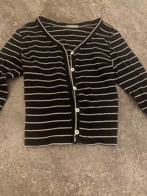 Svartvit randig kofta från Kappahl - Snygg svart kofta med vita ränder från Kappahl. Koftan har lång ärm, v-ringad hals och vita knappar framtill. Materialet är ribbad och känns mjukt mot huden. Perfekt för dig som gillar klassisk stil med en twist.