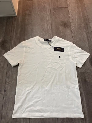 Vit t-shirt från Polo Ralph Lauren - Vit t-shirt från Polo Ralph Lauren med klassisk passform och korta ärmar. T-shirten har rund halsringning och den ikoniska broderade loggan på bröstet. Tillverkad i mjuk bomull för en skön känsla. Perfekt basic-plagg med stilren look.