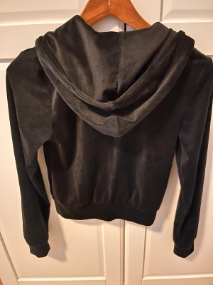 Svart velour hoodie från Gina Tricot - Svart hoodie i mjuk velour från Gina Tricot. Tröjan har huva, dragkedja med ringdetalj och två fickor fram. Långärmad och med ribbade muddar vid ärmslut och nederkant. Perfekt för chill dagar och snygg att matcha med jeans eller mjukisbyxor.