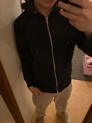 Svart hoodiejacka med dragkedja - Svart hoodiejacka med huva och snörning, hel dragkedja framtill och två fickor. Jackan har en enkel och clean look, perfekt för chill dagar. Passformen är normal och materialet känns mjukt och bekvämt mot kroppen.