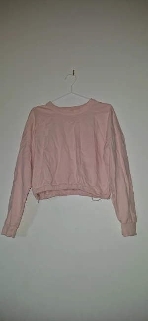 Croppad sweatshirt - Ljusrosa croppad sweatshirt från Etirel med långa ärmar och rund hals. Tröjan har snörning nertill för justerbar passform. Perfekt för en chill och avslappnad stil.