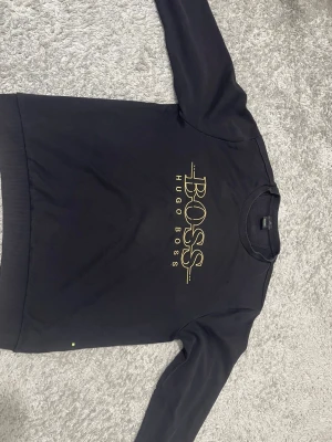 Svart Hugo Boss tröja med guldtryck - Svart långärmad tröja från Hugo Boss i slim fit-modell. Framsidan har ett stort BOSS-tryck i guld och rund halsringning. Materialet är mjukt och känns som bomull, perfekt för dig som gillar stilrena och lyxiga plagg med snygg logga. Den är XL men passar L.