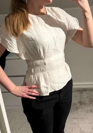 Vit blus  - Säljer en vit blus med snygg struktur och korta ärmar från H&M. Blusen har en markerad midja med smockdetaljer och en liten öppning i ryggen. Materialet är mjukt och luftigt, perfekt för vår och sommar. Passformen är normal och toppen är lätt att matcha.
