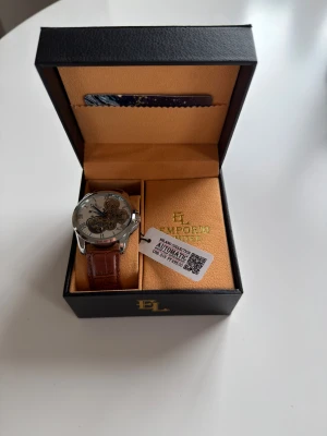 Emporio Limited automatisk klocka - Snygg automatisk klocka från Emporio Limited med transparent urtavla som visar mekaniken. Klockan har silverfärgat hölje, romerska siffror och silver visare. Armbandet är brunt och har en skinnlook med krokodilpräglad yta. Kommer i original ask.