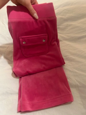 Juicy Couture mjukisset  - Rosa mjukisdress från Juicy Couture i mjuk velour med broderad JC-logga på fickan. Byxorna har bred resår i midjan och praktiska fickor, varav en med lock och silverfärgade knappar. Perfekta för chill eller streetwear. Tröjan är i stl M och byxorna stl S