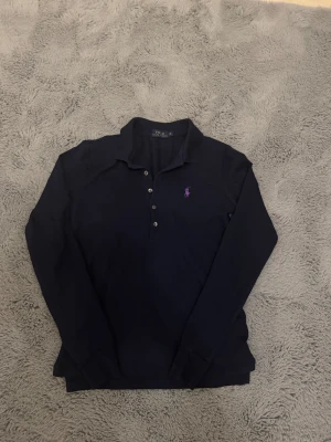 Mörkblå pikétröja Polo Ralph Lauren XS - Snygg mörkblå pikétröja från Polo Ralph Lauren i storlek XS. Klassisk krage, knappar framtill och lila broderad logga på bröstet. Tröjan är långärmad och gjord i mjuk bomull som är skön mot huden. Perfekt för dig som gillar stilrena och tidlösa plagg.