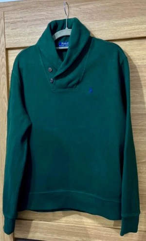 Grön långärmad tröja Polo Ralph Lauren - Snygg grön långärmad tröja från Polo Ralph Lauren med sjalkrage och knappar vid halsen. Tröjan har en broderad blå logga på bröstet och är tillverkad i mjuk bomull. Perfekt för dig som gillar klassisk stil med en twist. Sista bilden saxad för att visa exempel på kragen med skjorta under då det är dundersnyggt! 