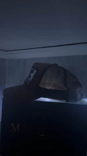 Brun Maggiore truckerkeps med mesh - Snygg brun truckerkeps från Maggiore med svart mesh på sidorna och bak. Framtill finns en stor patch med svartvit bild av personer och logga. Kepsen har böjd skärm och klassisk snapback-passform. Perfekt för dig som gillar streetstyle och vill sticka ut.