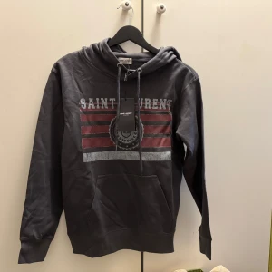 Grå Saint Laurent hoodie med tryck - Säljer en grå hoodie från Saint Laurent med coolt tryck i rött och vitt framtill. Tröjan har huva med snörning och en stor magficka. Perfekt för dig som gillar streetstyle och vill ha något snyggt och bekvämt.