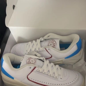 Nike Air Jordan 2 Low vit/blå/röd - Säljer ett par Nike Air Jordan 2 Low sneakers i vitt skinn med blå och röda detaljer. Klassisk siluett med perforerad tå, snörning och retro Air Jordan-logga på plösen. Perfekta för dig som gillar streetwear och vill sticka ut med en ikonisk modell.