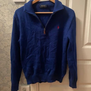 Blå stickad tröja Polo Ralph Lauren - Snygg blå stickad tröja från Polo Ralph Lauren med halv dragkedja och ribbad krage. Klassisk röd logga broderad på bröstet. Perfekt för lager-på-lager och passar till både jeans och chinos.
