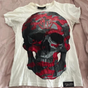 Vit t-shirt med dödskalle från Philipp Plein - Cool vit t-shirt från Philipp Plein med stor dödskalle framtill i röd, svart och grå camouflage. Klassisk passform och korta ärmar. T-shirten har en snygg logga nertill och är tillverkad i mjuk bomull. Väldigt  bra skick förutom att det är lite gult i kragen men inget man tänker på. 