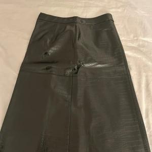 Svart långkjol i skinnlook från Visual Clothing Project - Snygg svart långkjol från Visualy Clothing Project med croco-präglad yta i skinnimitation. Kjolen har en stilren, rak passform och dragkedja i sidan. Perfekt för dig som gillar edgy och trendiga vibes.