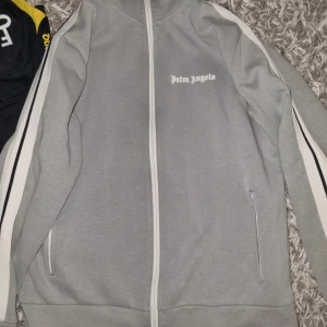 Grå Palm Angels zip hoodie med ränder - Snygg grå hoodie från Palm Angels med hel dragkedja och vita ränder längs ärmarna. Ikonisk logga på bröstet och praktiska fickor framtill. Perfekt för en chill och sportig look.