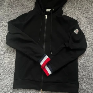 Svart Moncler zip up - Svart hoodie från Moncler med hel dragkedja, huva och snörning. Ärmsluten har snygga ränder i rött, vitt och blått. Moncler-logga på ena ärmen. Tillverkad i mjuk bomull, perfekt för en chill och stilren look. Helt ny och oanvänd med prislappen inuti. Byte funkar