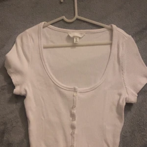 Vit croppad ribbad topp med knappar - Säljer en vit croppad topp från H&M med korta ärmar och djup rundringning. Toppen är ribbad och har knappar framtill som en snygg detalj. Perfekt för dig som gillar en enkel men trendig stil.