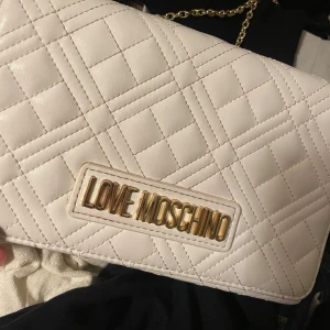 Vit quiltad axelväska Love Moschino - Supersnygg vit quiltad axelväska från Love Moschino med guldfärgad kedja och logga framtill. Väskan har ett elegant rutmönster och stängs med magnetknapp. Perfekt för dig som vill ha en stilren och trendig accessoar.