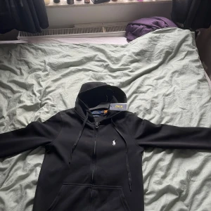 Svart hoodie från Polo Ralph Lauren - Svart hoodie från Polo Ralph Lauren med vit broderad logga på bröstet. Tröjan har dragkedja, huva med snörning och känguruficka. Tillverkad i mjuk bomullsmix och har en clean, klassisk look som funkar till allt.