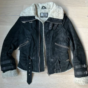 Jofama by Kenza jacka  - Snygg svart teddyjacka från Jofama by Kenza med mjukt beige foder, bälte i midjan och detaljerade spännen vid ärmslut och krage. Jackan har dragkedjor på fickor och framsida samt coola metalldetaljer. Perfekt för dig som vill ha en statementjacka med edge. 