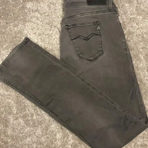 Replay grå slimfit jeans med slitningar och Hyperflex  - Snygga grå jeans från Replay med slim fit och slitna detaljer på benen. Klassisk femficksmodell med svarta sömmar och Replay-logga på bakfickan. Jeansen är i stretchigt material för en tight passform och har knäppning med knapp och dragkedja.
