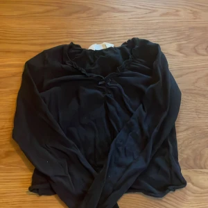 Svart blus med volang och rosett H&M - Svart blus från H&M med lång ärm, volangdetalj längs ringningen och en liten söt rosett framtill. Tunn och lätt i materialet, perfekt för dig som gillar en stilren men ändå gullig look.