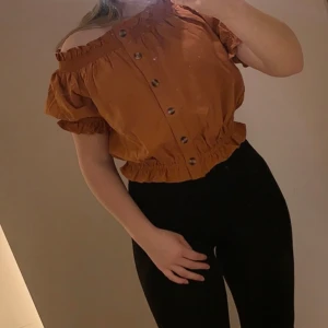 Offshoulder blus med knappar - Säljer en snygg senapsgul/orange blus med offshoulder-modell och korta puffärmar. Toppen har knappar framtill, resår vid halsringning, ärmslut och midja samt en croppad passform. Perfekt för dig som gillar trendiga och bekväma plagg med lite volym. Mycket fint skick!