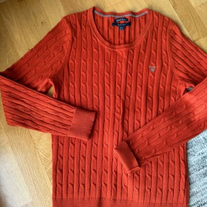 Orange kabelstickad tröja från GANT - Kabelstickad orange tröja från GANT. Klassisk rund halsringning, ribbade muddar och diskret logga broderad på bröstet. 