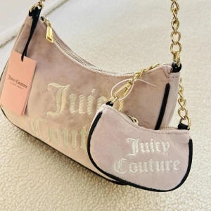 Juicy Couture rosa axelväska i sammet - Snygg rosa axelväska från Juicy Couture i mjuk sammet med matchande mini bag. Väskan har guldfärgade kedjedetaljer, svart kant och broderad logga framtill. Perfekt för dig som vill sticka ut med en trendig accessoar.