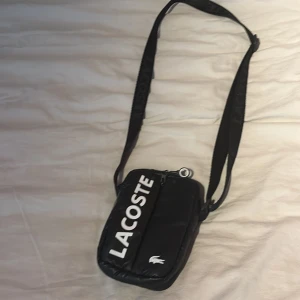 Svart axelväska från Lacoste - Snygg svart axelväska från Lacoste med vit logga framtill och justerbar rem. Väskan har dragkedja och orange foder på insidan samt meshficka. Perfekt storlek för mobil, plånbok och andra småprylar.