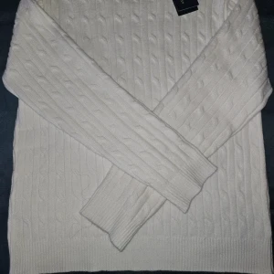 Vit kabelstickad tröja Polo Ralph Lauren - Vit kabelstickad tröja från Polo Ralph Lauren med klassisk rund halsringning och broderad logga på bröstet. Tröjan har långa ärmar och ribbade muddar för en snygg och tidlös look. Perfekt för dig som gillar stilrena och exklusiva plagg.