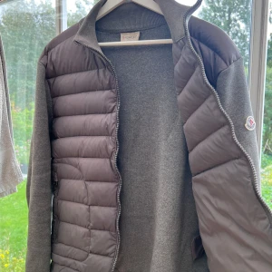 Grå Moncler cardigan med stickade ärmar - Snygg grå cardigan från Moncler med quiltad front och stickade ärmar. Jackan har dragkedja framtill och två fickor med dragkedja. Klassisk Moncler-logga på vänster ärm. Perfekt för höst och vinter när du vill ha både stil och komfort. Pris går att diskutera