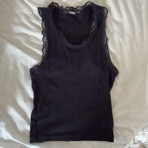 Svart spetskantad linnetopp Bik Bok - Svart ärmlös linnetopp från Bik Bok med fina spetsdetaljer längs halsringning och ärmhål. Toppens enkla design gör den lätt att matcha och den har en mjuk bomullskänsla. Perfekt för att skapa en stilren look.