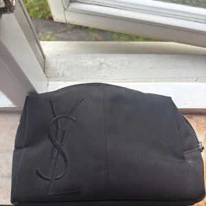 Svart necessär från Yves Saint Laurent - Snygg svart necessär från Yves Saint Laurent med stort YSL-logo broderat på framsidan. Väskan har en enkel, stilren design och stängs med dragkedja. Perfekt för att organisera smink eller småprylar. Tillverkad i slitstark polyester.