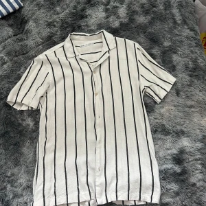 Randig kortärmad skjorta från Zara - Vit skjorta från Zara med svarta vertikala ränder, klassisk krage och knäppning framtill. Tillverkad i luftig linnemix, perfekt för varma dagar. Skjortan har korta ärmar och en avslappnad passform som ger en chill vibe.