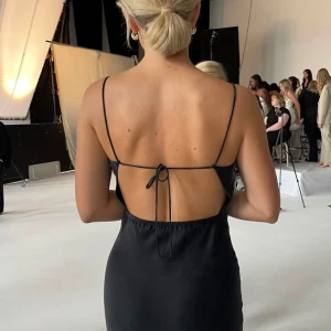 Svart långklänning med öppen rygg - Supersnygg svart långklänning med tunna axelband och öppen rygg med snörning. Klänningen har en elegant, figurnära passform och är tillverkad i ett glansigt, mjukt material som ger en lyxig känsla. Perfekt för dig som vill sticka ut med en stilren och modern look.